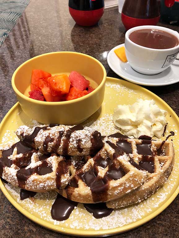 chocolate covered waffle with bowl of fruit and hot cocoa at breakfast table