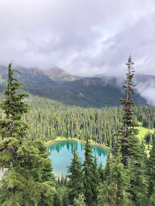 teal circular lake surrounded by forest trees and clouds in the distance