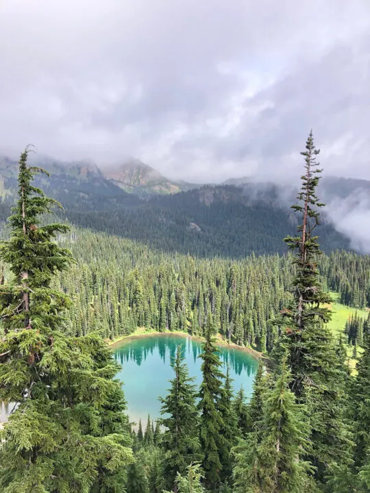 teal circular lake surrounded by forest trees and clouds in the distance