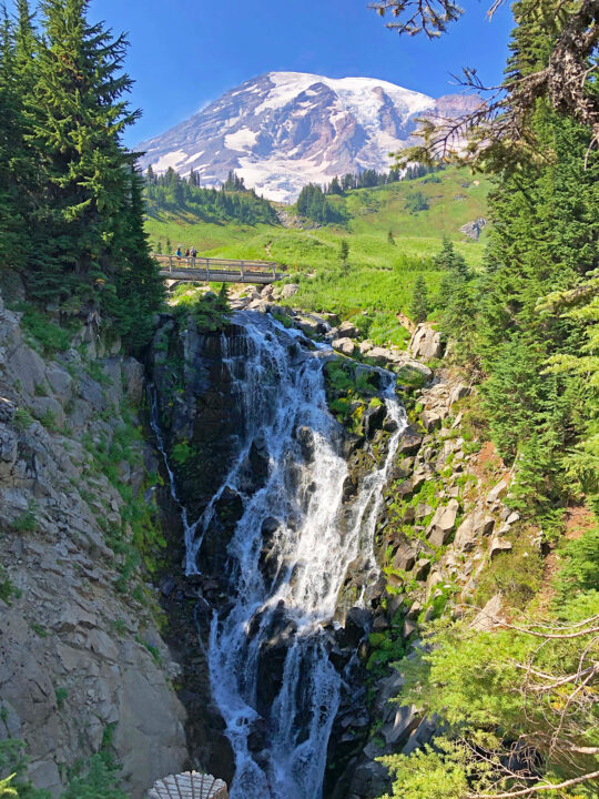 beautiful waterfall running down cliff surrounded by trees and mountain peak in distance view while on the best hikes in mt rainier