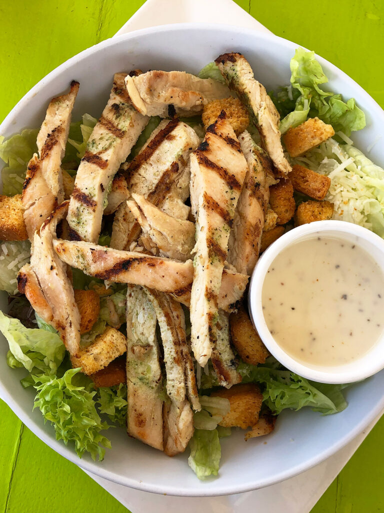 chicken cesar salad with strips of chicken cup of dressing in white bowl on green table