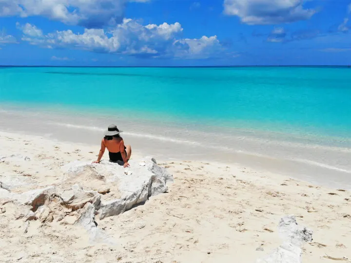 cheapest island vacations picture of woman sitting on rock with white sandy beach and teal water with blue sky puffy clouds