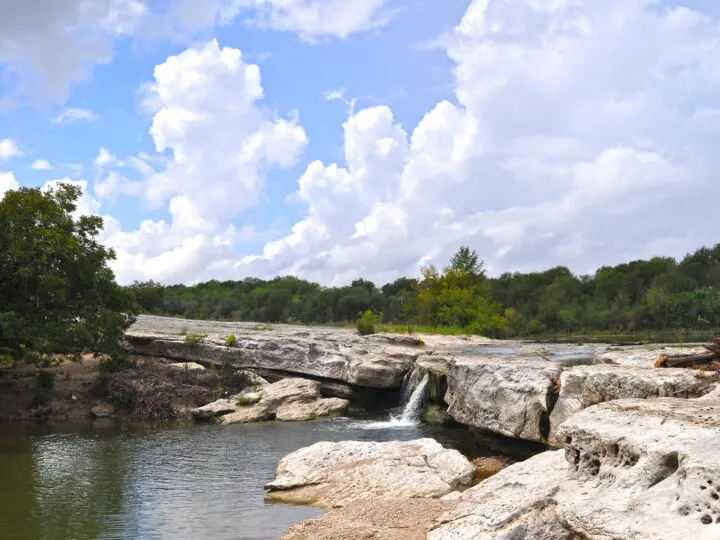 hiking in Austin photo of lake with small waterfall large rocky cliff with trees on cloudy day