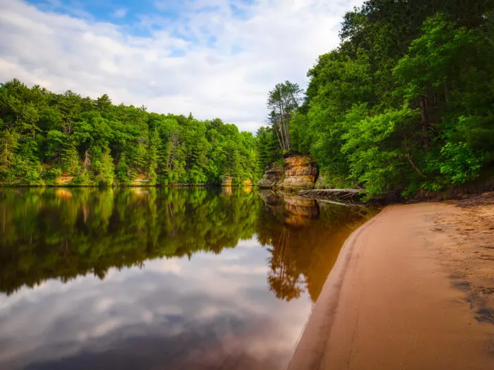 midwest road trip photo of lake with tan sand trees and reflection of cloudy sky