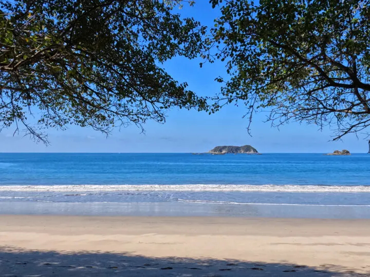 picture of tan sandy beach in Manuel Antonio with blue water and trees framing the top portion of photo