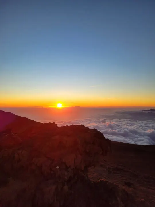 view of sun dipping into horizon orange with grey clouds and rocks in foreground