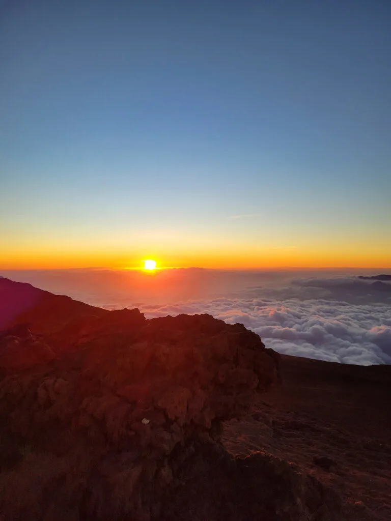 view of sun dipping into horizon orange with grey clouds and rocks in foreground