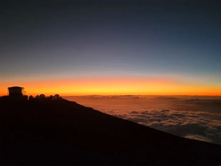 mountainside in dark with sunset in distance above the clouds
