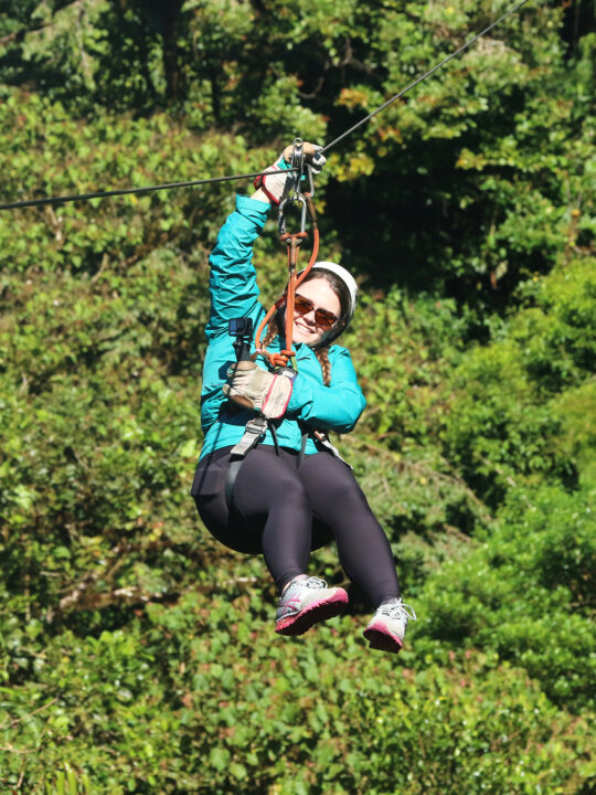 woman zip lining in jungle with black pants teal jacket