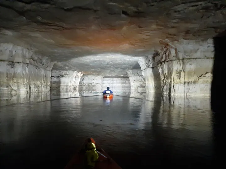 adventure vacations in the US: view of inside of cave from kayak dark water grey and orange cave and spotlight on kayaker ahead