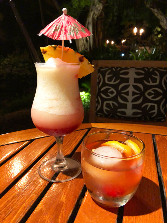 tall glass with fruity drink and umbrella small cocktail glass on wood table at one of the best places to eat in kauai