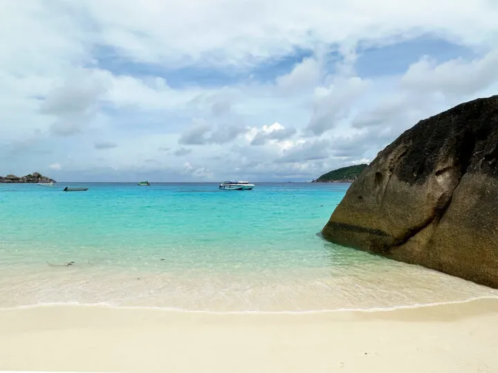 white sand beach in thailand with large rock on side blue water clouds in sky