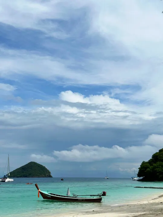 Koh Hey Thailand view of beach and boats in teal water with island in distance clouds in sky