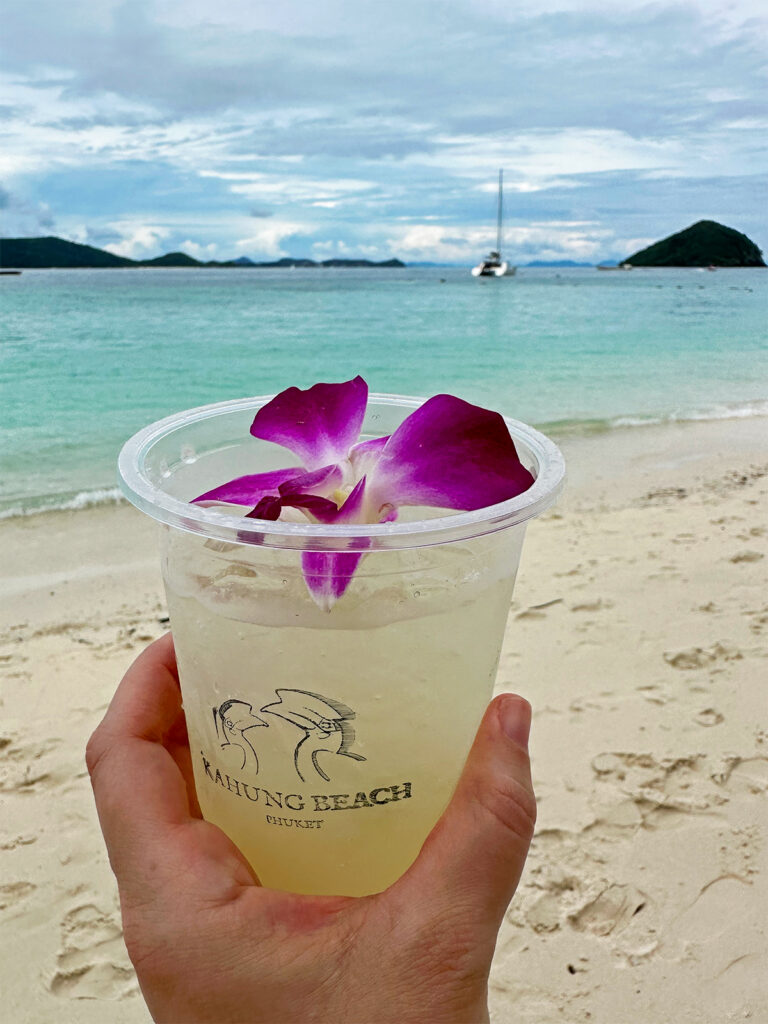 hand holding margarita with purple flower with beach in background coral island review