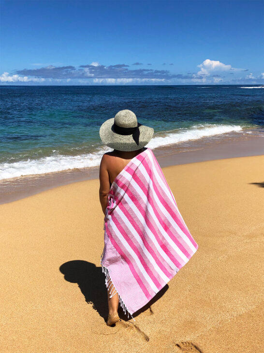 what to pack for hawaii view of woman standing on beach in hat and striped pink towel