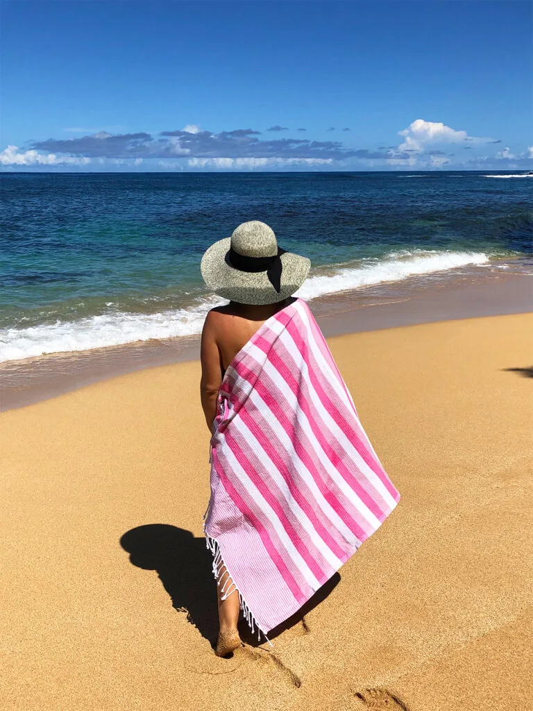 view of woman standing on beach in hat and striped pink towel