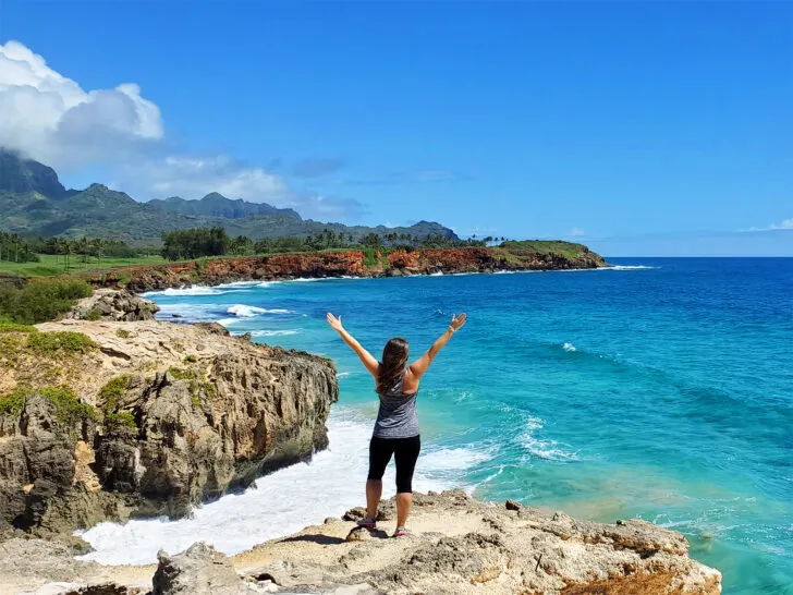 woman standing at edge of cliff in kauai with arms up high with blue ocean