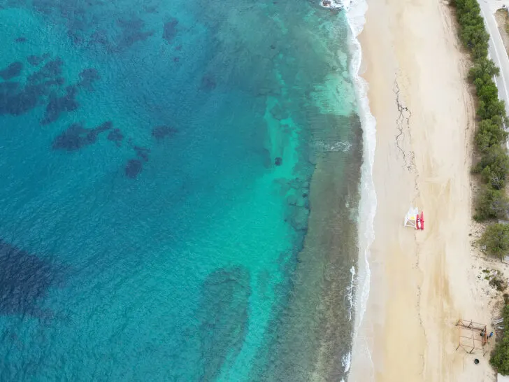 Mykonos vs. Santorini view of Kalafati Beach in Mykonos from above with tan sand white ocean wave and blue and teal sea water