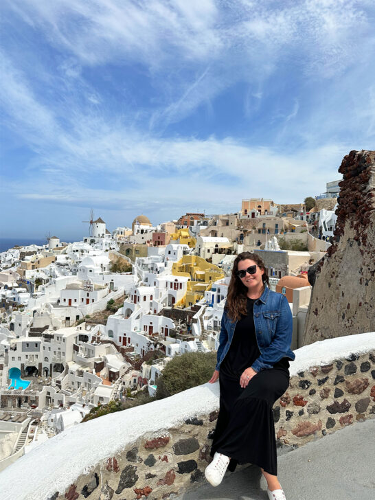woman wearing black dress and jean jacket with white shoes and white buildings in background battle of Mykonos vs Santorini