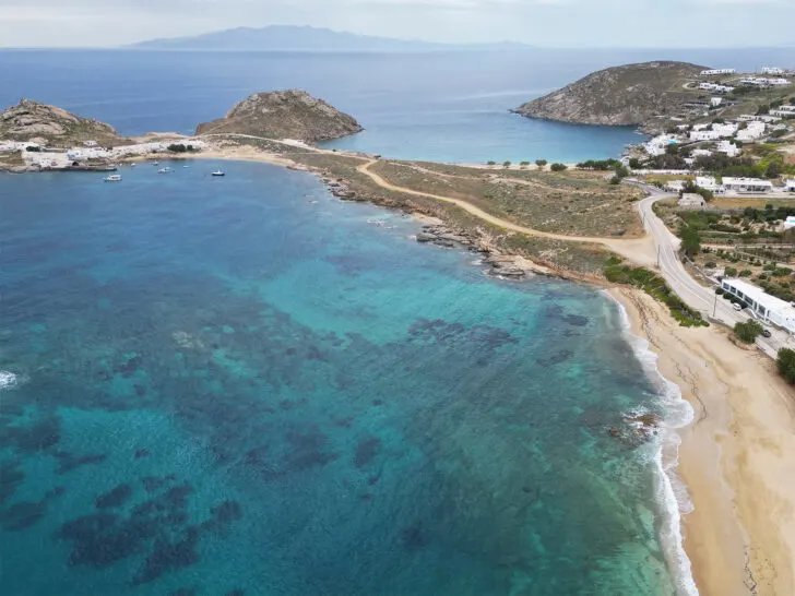 aerial view of beach with blue and teal water with hilly coastline Santorini vs Mykonos