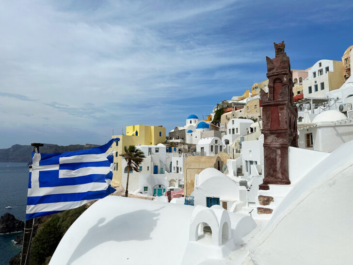 Santorini vs. Mykonos view of hillside village with Greece flag and colorful buildings blue sky with clouds