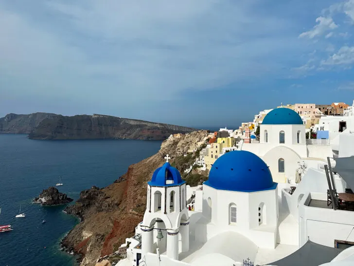 Mykonos vs Santorini view of Santorini blue dome church and town in distance atop huge hill with ocean below