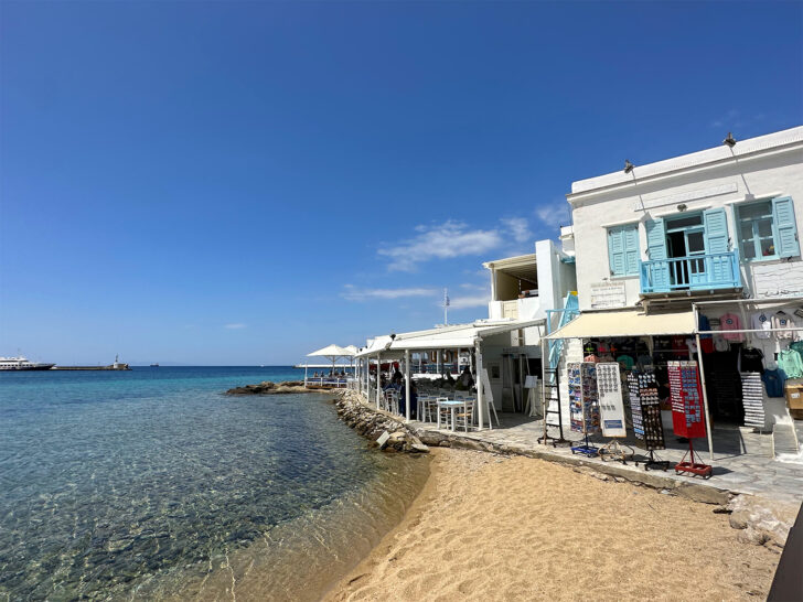 shops lining waterfront with small beach in Mykonos Greece