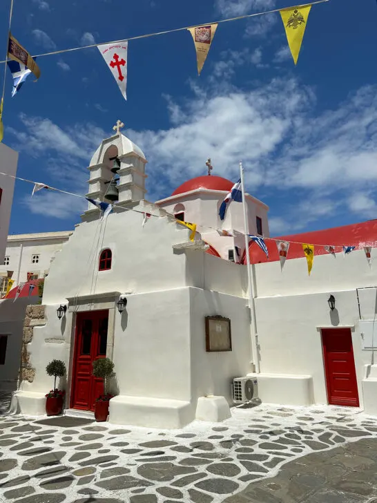 Mykonos vs Santorini view of church with white and red on stone streets with flags above