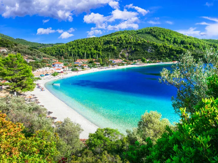 couples holiday to greece view of bright blue water, white sand and hillside with hotel buildings