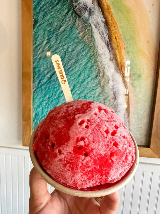 hand holding red shave ice with spoon that says Ululani's with poster behind