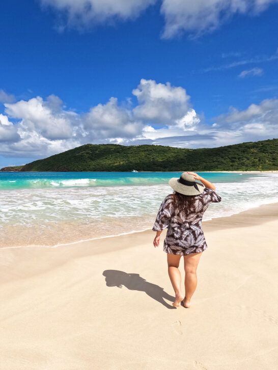 woman walking with hand on hat on white sand beach with blue water and hilly coastline in distance on the beach