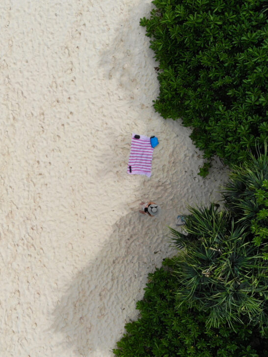 woman standing in shade of trees with beach towel on sand while flying a drone