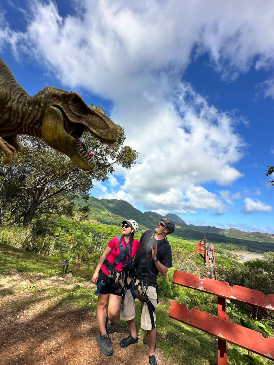 man and woman in zipline attire with fake dinosaur on screen in Hawaii