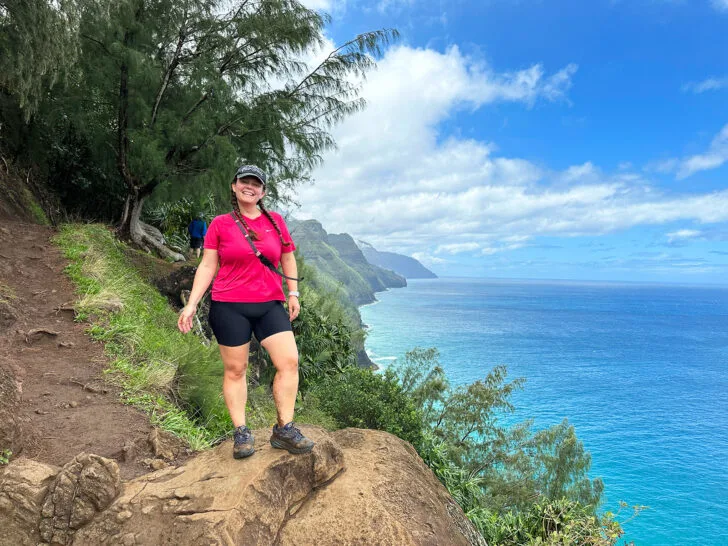 woman in pink shirt and black shorts standing on edge of cliff with ocean in distance