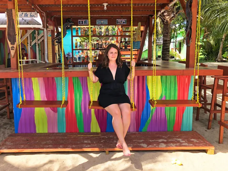 what to pack for a beach vacation woman sitting on swing on beach with multicolored bar behind wearing black dress