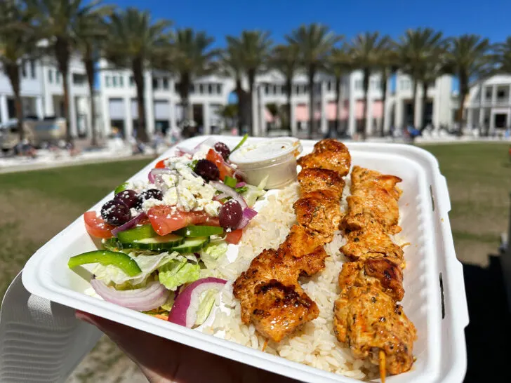 greek salad and kebabs in box with palm trees in the distance