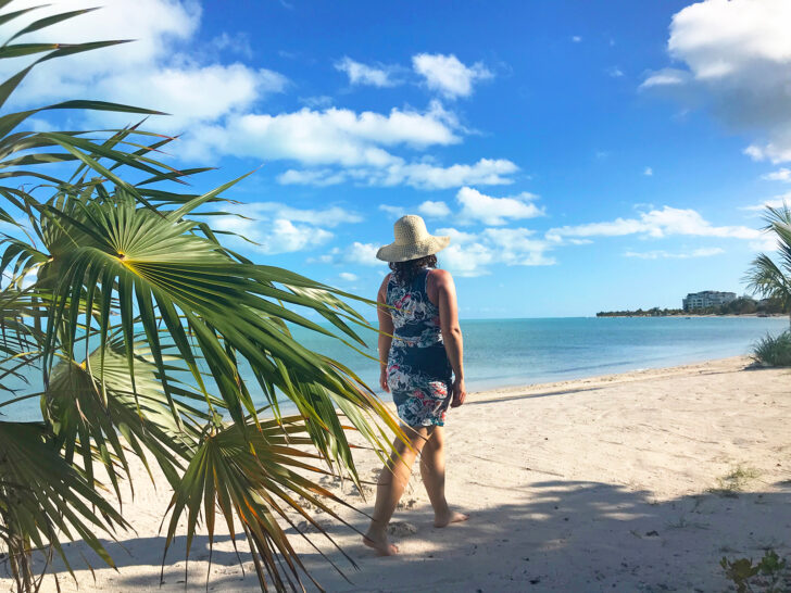 woman walking on beach in swim coverup with palm tree and hat on