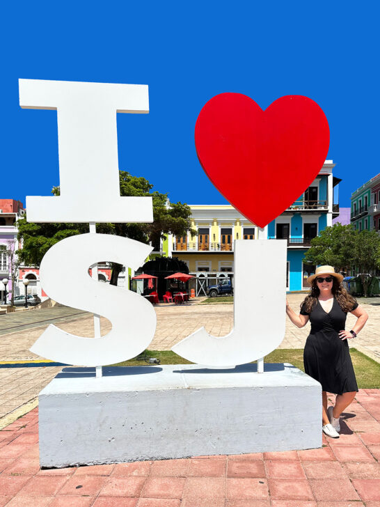 woman standing to the right of a giant I heart SJ sign with colorful buildings in the background