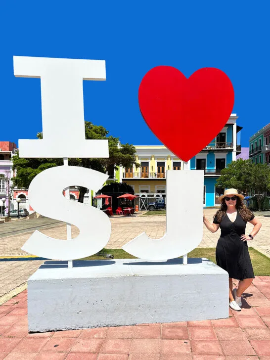 woman standing to the right of a giant I heart SJ sign with colorful buildings in the background