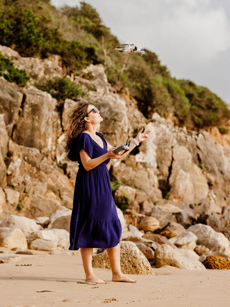 woman catching drone in the air wearing blue dress with rocky shoreline in distance