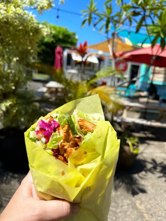 hand holding wrap with chicken and lettuce with food truck park in distance