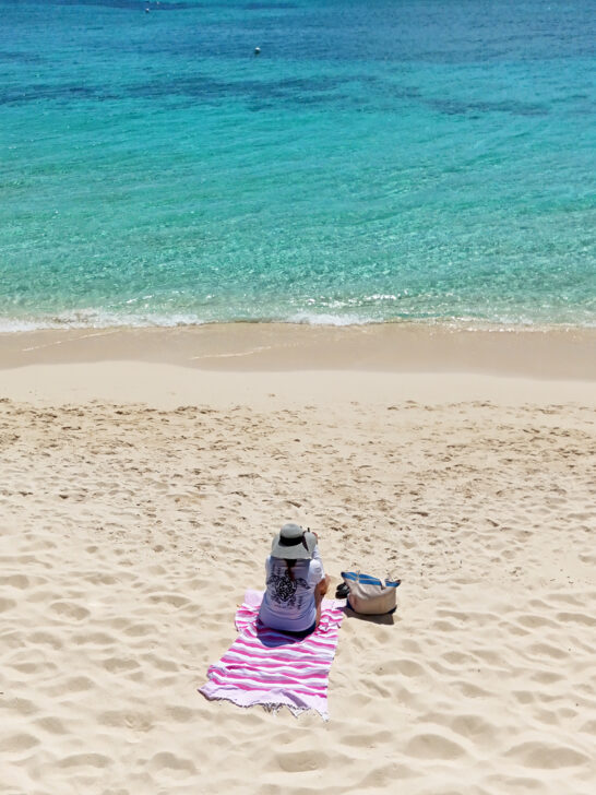 woman sitting on pink towel with white sand and teal water