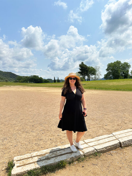 woman standing in black dress and hat on original olympic starting line stones with dirt track behind on a Greece mainland itinerary