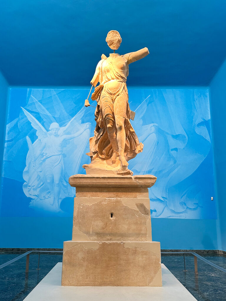 view of stone statue of Nike on podium with blue walls and ceiling behind in Ancient Olympia