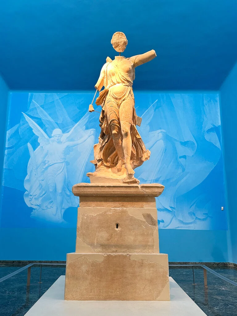 view of stone statue of Nike on podium with blue walls and ceiling behind in Ancient Olympia